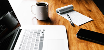 木桌上放著筆電、筆記、筆、手機與白色馬克杯裝的黑咖啡